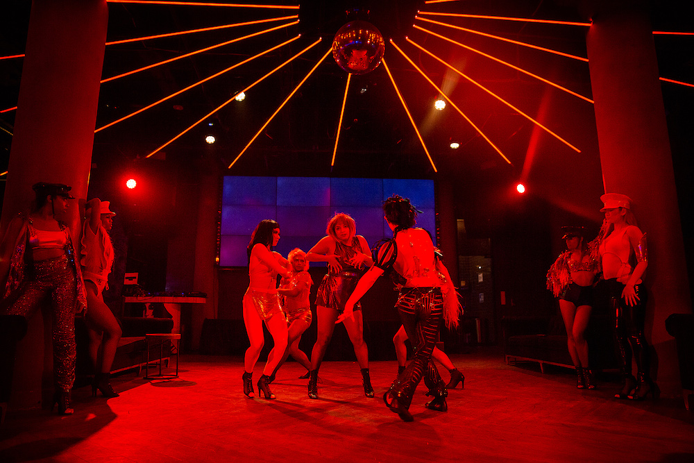 bathed in red light, a circle of club dancers focus on each other in ecstatic dancing as they are watched intently by people leaning on the columns of the club's interior